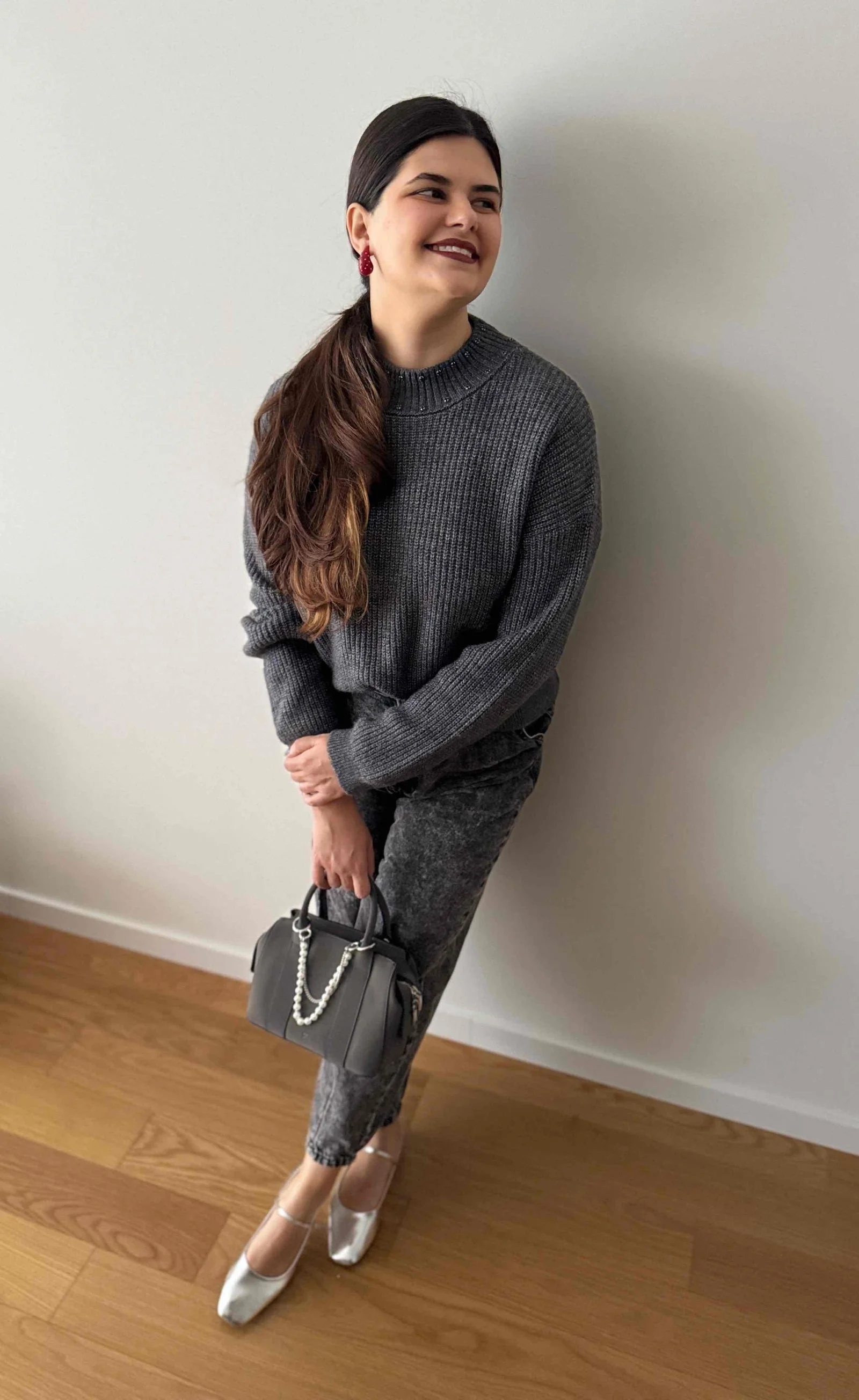 Francy sorrindo, usando um suéter cinzento e calças cinzentas, segurando uma bolsa preta, com brincos vermelhos